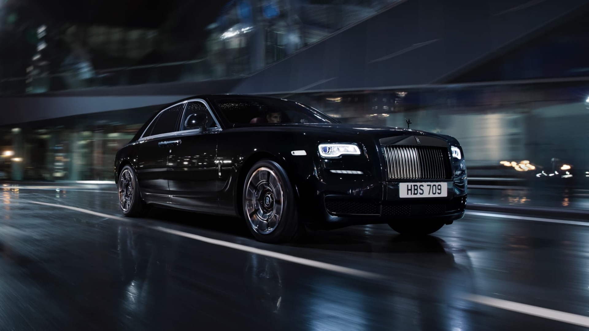 A dynamic shot of a Rolls-Royce Black Badge Wraith driving with speed in a dark cityscape in the rain